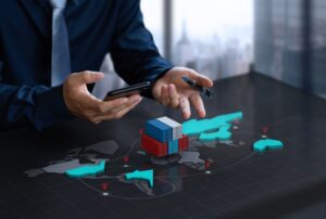 A businessman in a dark suit holding a smartphone and pen, analyzing a 3D holographic map with shipping containers and global trade routes.