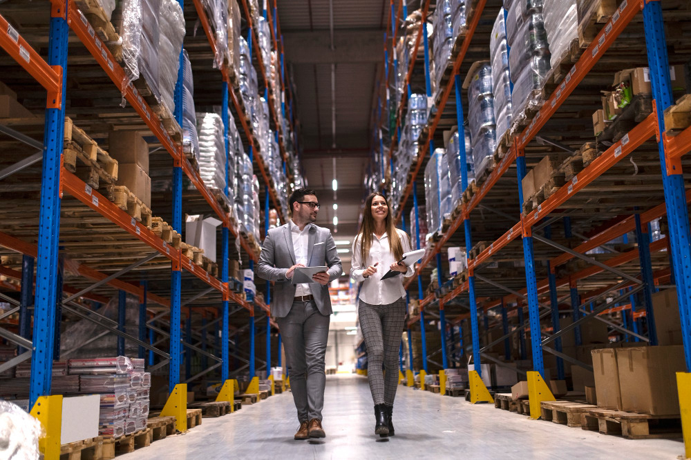 Two supply chain professionals walking through a warehouse aisle, inspecting inventory and discussing logistics operations.