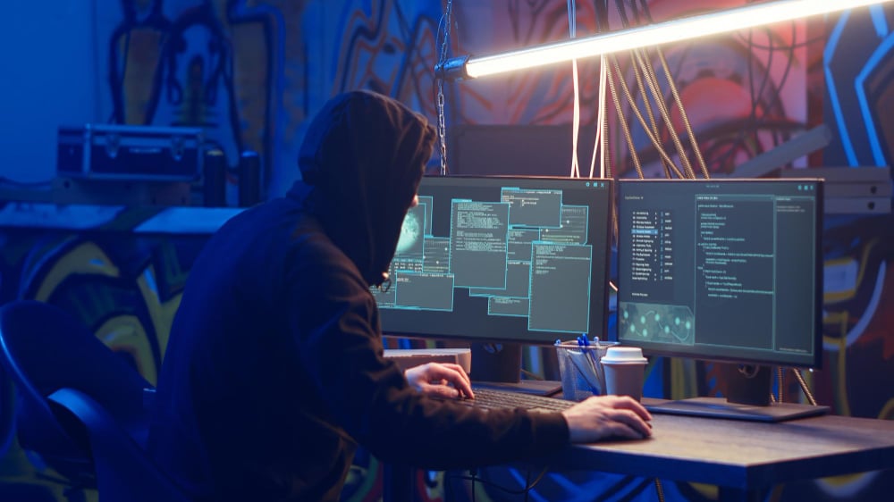 Hacker in a dark hoodie working on multiple computer screens in a dimly lit room, representing a supply chain cyber attack scenario